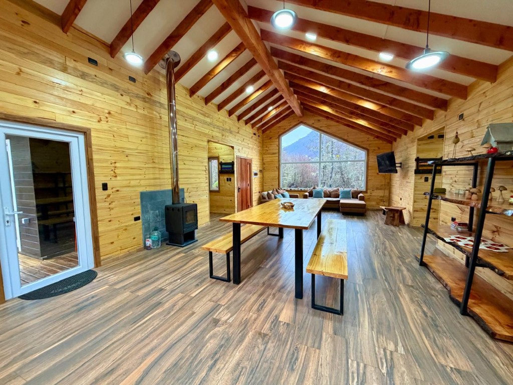 Living y comedor de Refugio Kürü desde el acceso, con estufa, mesas de madera y ventanal al fondo