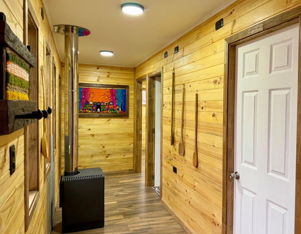Pasillo de madera de Refugio Kürü con estufa a leña, tejidos y accesos a las habitaciones